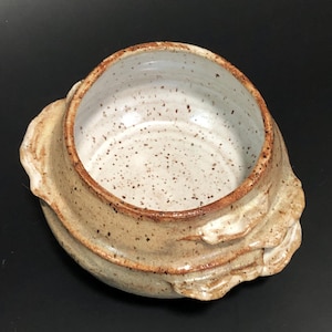 Op de afbeelding: Een handgemaakte keramische kom met een gespikkeld wit interieur en een getextureerde, gelaagde buitenkant in tinten bruin en bruin. De rand heeft een donkerbruin accent. De kom staat tegen een zwarte achtergrond.