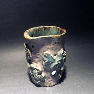 Op de afbeelding: Een kleine, cilindrische keramische vaas met een getextureerd, gevlekt bruin en groen glazuur. De vaas heeft een uniek ontwerp met uitgesneden bladvormen aan de zijkanten.