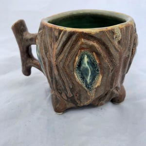 Pode incluir: Uma caneca de cerâmica artesanal com um design de tronco de árvore. A caneca apresenta um exterior castanho com detalhes texturizados semelhantes à casca, um interior verde e uma pega em forma de ramo. A caneca é suportada por três pequenos pés.