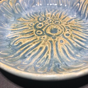 May include: A handmade ceramic bowl with a floral design. The bowl features a light blue glaze with a contrasting tan-colored floral pattern in the center. The bowl's rim is a lighter shade of blue.