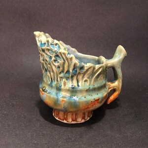 Op de afbeelding: Een handgemaakte keramische creamer met een uniek ontwerp. De creamer heeft een getextureerd, organisch patroon in tinten blauw, groen en bruin. Het heeft een handvat en een decoratieve basis. De creamer is ongeveer 10 cm hoog.