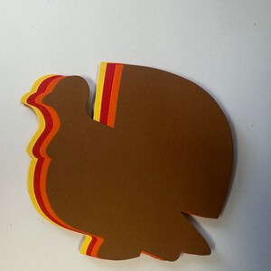 May include: A layered paper craft turkey cutout in shades of brown, orange, and yellow. The turkey is facing left and has a rounded body and a long, curved neck.
