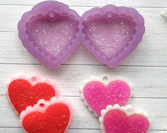 May include: An assortment of heart-shaped resin crafts in various stages. Includes a mold, finished red and pink hearts, and a sign that reads "LA.Charmed" with floral accents. The background is a white wooden surface.
