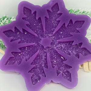 Puede incluir: Adorno morado en forma de copo de nieve con purpurina incrustada en el centro. El copo de nieve tiene cortes detallados y una estrella en el centro. El adorno probablemente está hecho de resina o un material similar.