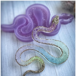 Puede incluir: Una figura decorativa de serpiente hecha a mano con un degradado de colores azul, verde y morado, y una cabeza y borde dorados. Un molde morado con forma de serpiente está en el fondo. La figura está sobre una superficie de madera azul claro.
