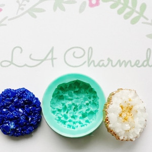 May include: An image featuring a blue crystal, a teal silicone mold, and a white and gold geode. The mold is round with a textured interior. The geode has a gold-colored edge. The background has a floral design and the text "L A Charmed".
