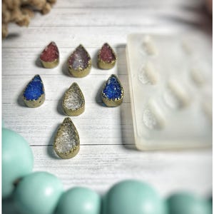 May include: A collection of teardrop-shaped pendants with gold-colored edges and colorful crystal-like centers in red, purple, blue, and clear. A silicone mold with teardrop shapes is visible, along with a strand of light blue beads.