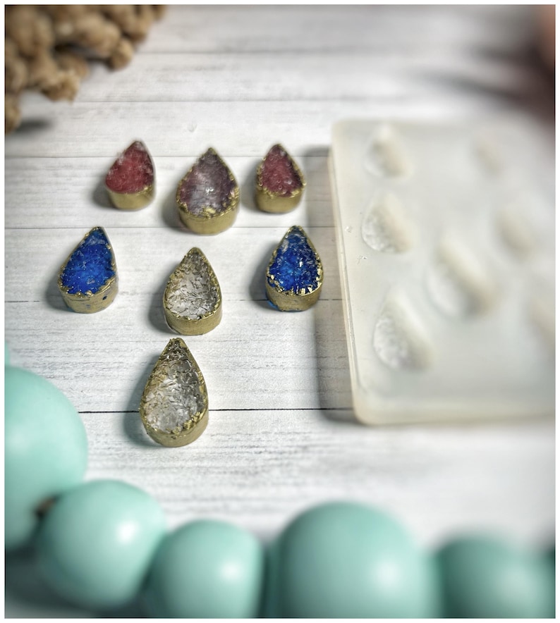 May include: A collection of teardrop-shaped pendants with gold-colored edges and colorful crystal-like centers in red, purple, blue, and clear. A silicone mold with teardrop shapes is visible, along with a strand of light blue beads.