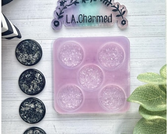 May include: A collection of crafting supplies, including a lavender-colored silicone mold with five circular cavities, and four black resin cabochons with silver glitter. The image also shows a sign that says "LA.Charmed".