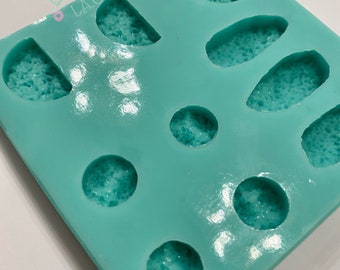 May include: A turquoise silicone mold with various shapes, including half-circles, ovals, and circles. The mold is filled with a textured, light blue substance. The text "LA.Charmed" is visible in the upper left corner.
