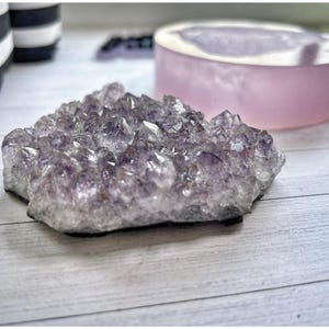 Peut inclure: Un amas de cristaux d'améthyste, présentant diverses nuances de violet. Les cristaux sont disposés de manière naturelle et irrégulière. Un objet rond rose clair est en arrière-plan. Les cristaux sont sur une surface en bois blanc.