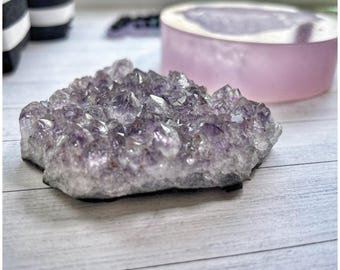 May include: A cluster of amethyst crystals, showcasing a range of purple hues. The crystals are arranged in a natural, irregular formation. A light pink, round object is in the background. The crystals are on a white wooden surface.