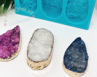 May include: A teal silicone mold with three teardrop-shaped cavities, alongside three gemstone pendants in purple, white, and blue. Each pendant has a gold-colored edge. The pendants are on a white surface.