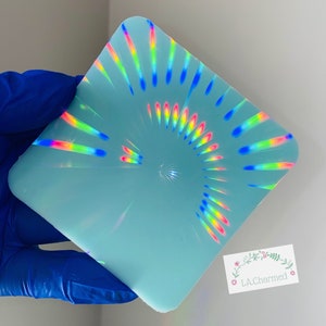 Puede incluir: Una pieza de resina azul claro, de forma cuadrada, con un diseño holográfico de arcoíris. El diseño irradia desde el centro, creando un efecto colorido y llamativo. Se ve una esquina de una etiqueta blanca con el texto "LA.Charmed".