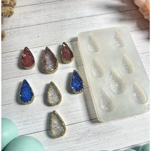 May include: A collection of teardrop-shaped resin pendants in various colors, including red, purple, blue, and clear, with gold-colored edges. A silicone mold with matching teardrop shapes is also present, suggesting a crafting theme.