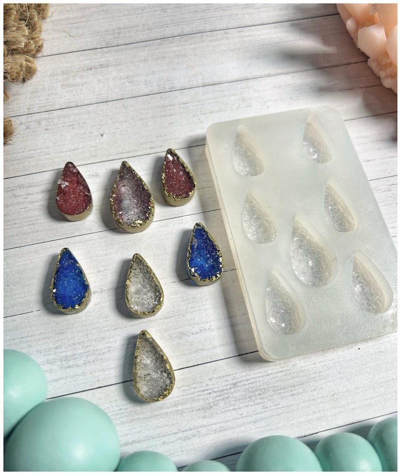 May include: A collection of teardrop-shaped resin pendants in various colors, including red, purple, blue, and clear, with gold-colored edges. A silicone mold with matching teardrop shapes is also present, suggesting a crafting theme.
