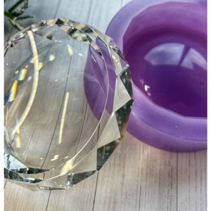 May include: A clear, geometric crystal paperweight with a faceted design. Next to it is a purple, round silicone mould. The paperweight reflects light, creating a rainbow effect. The items are on a light-coloured wooden surface.
