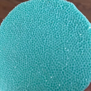 May include: A close-up of a round, light blue object with a textured surface. The surface is covered in small, evenly spaced holes, giving it a porous appearance. Small, shiny particles are scattered across the surface.