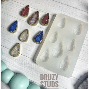 May include: A collection of teardrop-shaped druzy studs in various colors, including red, blue, and silver, with gold-colored edges. A silicone mold with matching teardrop shapes is also present, along with the text "DRUZY STUDS".