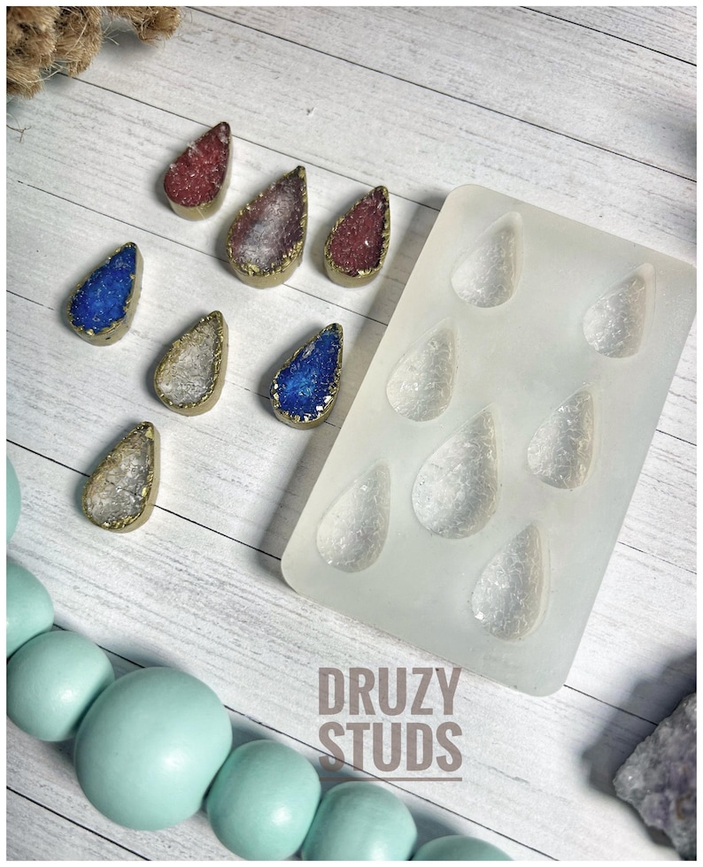 May include: A collection of teardrop-shaped druzy studs in various colors, including red, blue, and silver, with gold-colored edges. A silicone mold with matching teardrop shapes is also present, along with the text "DRUZY STUDS".