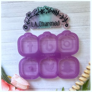 Pode incluir: Um molde de silicone roxo com ícones de mídia social: Facebook, TikTok, Instagram, YouTube, Twitter e Snapchat. O molde tem o texto "L.A. Charmed". Um design floral decorativo está acima.