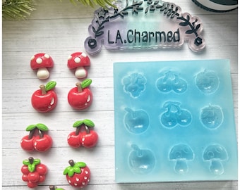 Puede incluir: Una colección de dijes de resina con temática de frutas y un molde de silicona. Los dijes incluyen manzanas rojas, cerezas, fresas y champiñones con puntos blancos. El molde es azul y presenta formas de frutas y champiñones. El texto "LA.Charmed" es visible.