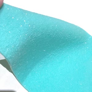 Könnte beinhalten: Nahaufnahme eines Stücks türkisfarbenen Materials mit strukturierter Oberfläche und verstreutem Glitzer. Das Material ist leicht gebogen und gibt einen Blick auf einen weißen Hintergrund frei. Das Gesamtbild deutet auf ein dekoratives oder handwerkliches Element hin.