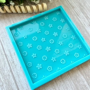 Può includere: Uno stampo quadrato in resina blu chiaro con un motivo ripetuto di fiori e stelle in rilievo. Lo stampo ha un bordo rialzato ed è progettato per l'artigianato. Lo sfondo è una venatura del legno chiaro.