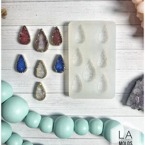 May include: A collection of teardrop-shaped resin pendants in red, blue, and clear, with gold-colored edges. A silicone mold with matching teardrop shapes is also present. The pendants are displayed on a white wooden surface, with a light blue beaded strand.