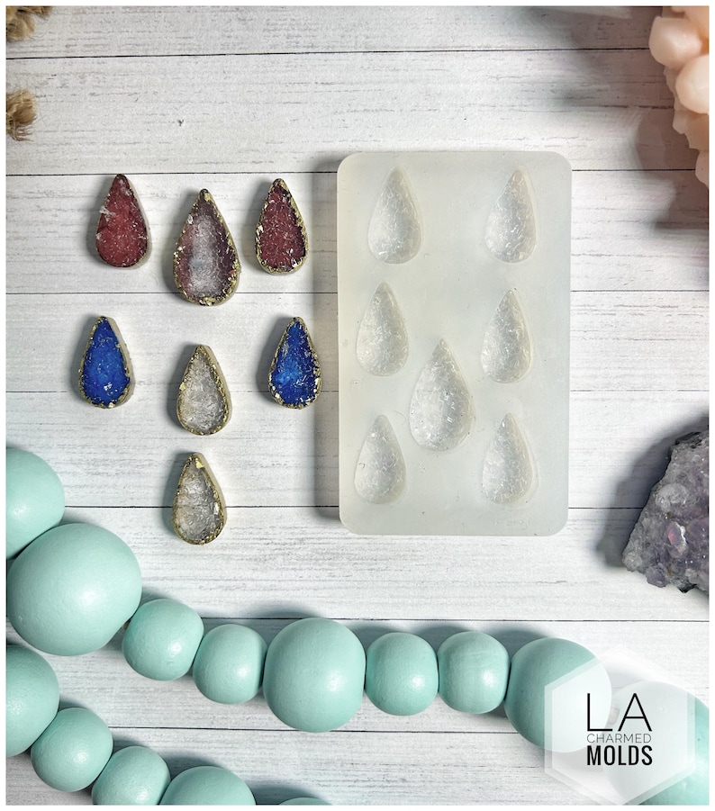 May include: A collection of teardrop-shaped resin pendants in red, blue, and clear, with gold-colored edges. A silicone mold with matching teardrop shapes is also present. The pendants are displayed on a white wooden surface, with a light blue beaded strand.