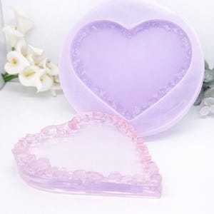 May include: Two heart-shaped moulds, one large and one small, in shades of lavender and pink. The moulds have a textured border, likely for resin crafts. The background is white.