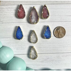 May include: A collection of teardrop-shaped pendants with gold-colored edges and colorful, textured centers. The pendants are in shades of red, purple, blue, and clear. A penny is included for size reference. The pendants are arranged on a white wooden surface.