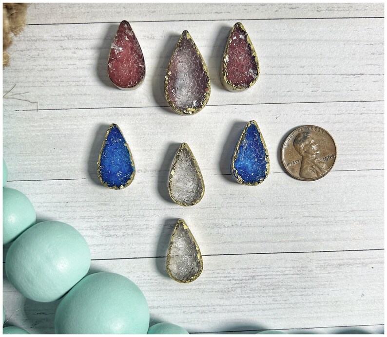 May include: A collection of teardrop-shaped pendants with gold-colored edges and colorful, textured centers. The pendants are in shades of red, purple, blue, and clear. A penny is included for size reference. The pendants are arranged on a white wooden surface.
