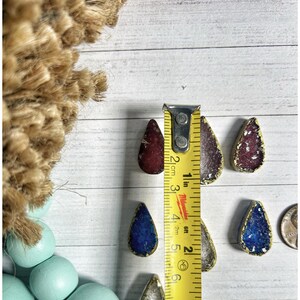May include: A collection of teardrop-shaped pendants in various colors, including red, purple, blue, and silver, with gold-colored edges. The pendants are approximately 1 inch in length. A yellow measuring tape is placed next to the pendants.