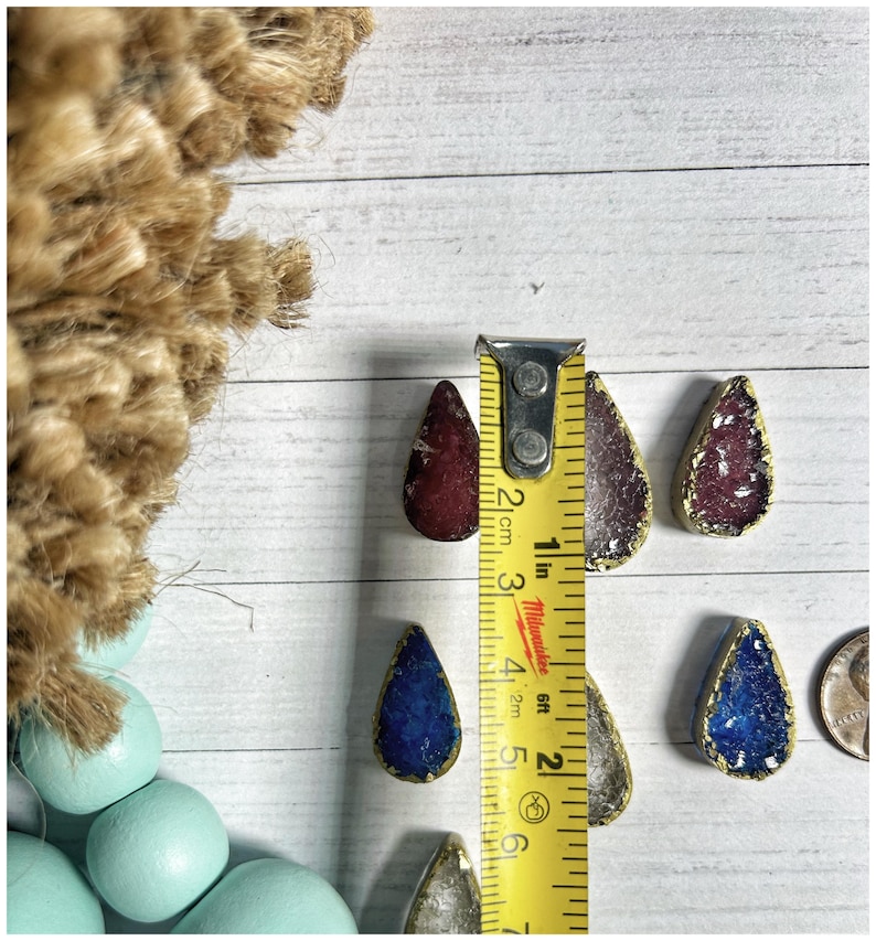 May include: A collection of teardrop-shaped pendants in various colors, including red, purple, blue, and silver, with gold-colored edges. The pendants are approximately 1 inch in length. A yellow measuring tape is placed next to the pendants.