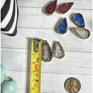 May include: Teardrop-shaped pendants with gold-colored edges and various colored interiors, including red, blue, purple, and clear. A yellow measuring tape shows the pendants are approximately 1 inch long. A penny is also in the image.