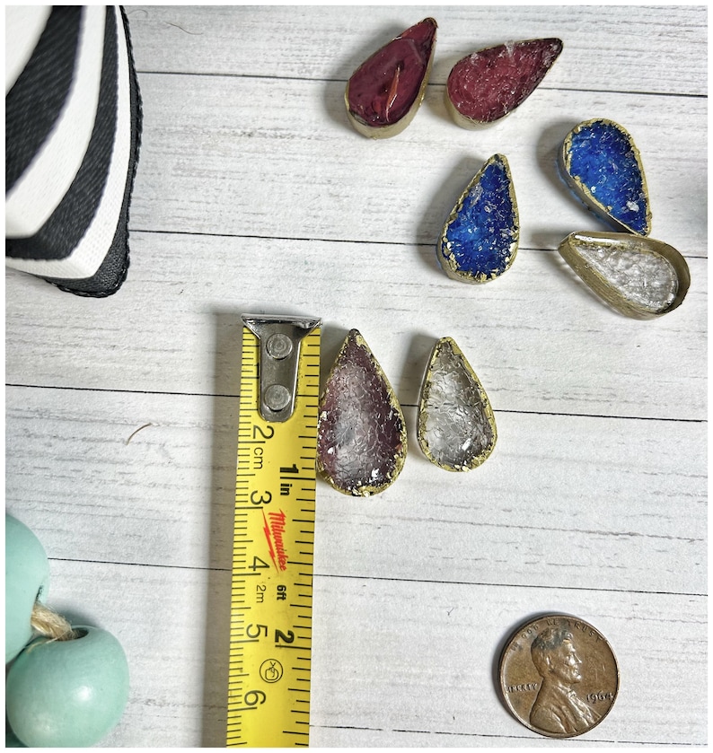 May include: Teardrop-shaped pendants with gold-colored edges and various colored interiors, including red, blue, purple, and clear. A yellow measuring tape shows the pendants are approximately 1 inch long. A penny is also in the image.