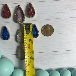 May include: A collection of teardrop-shaped pendants with gold edges and colorful, textured centers in red, blue, and clear. A ruler shows the pendants are approximately 1 inch in length. A penny is also included for size reference.