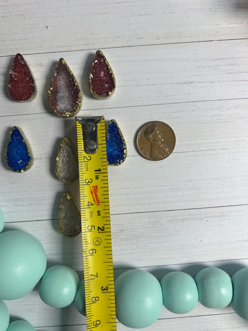May include: A collection of teardrop-shaped pendants with gold edges and colorful, textured centers in red, blue, and clear. A ruler shows the pendants are approximately 1 inch in length. A penny is also included for size reference.