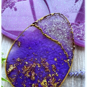Puede incluir: Un posavasos artesanal de resina morada con detalles de pan de oro y borde dorado. El posavasos tiene una forma orgánica única y está sobre una superficie de vetas de madera clara. Un molde en forma de corazón a juego es visible en el fondo.