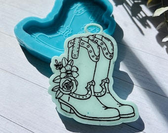May include: A light blue silicone mold in the shape of a cowboy boot, with a matching boot-shaped resin casting. The resin boot is outlined in black and decorated with flowers. The mold is on a white surface.