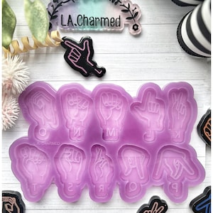 Puede incluir: Un molde de silicona lavanda con gestos de manos para letras y un dije de gesto de mano negro y rosa. También es visible un diseño floral con el texto "LA.Charmed". La imagen está sobre una superficie de madera blanca.