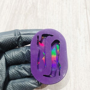 May include: A purple silicone mould in an oval shape, featuring two high-heeled boot outlines. The boots have a holographic, iridescent appearance. The mould is held by a gloved hand, with a light wood-grain surface in the background.