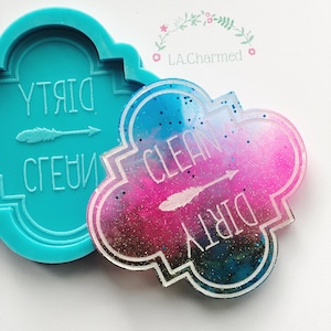 Puede incluir: Un molde de resina turquesa y un letrero de resina terminado. El letrero terminado tiene un borde blanco y presenta las palabras "CLEAN" y "DIRTY" con una flecha. El letrero tiene un diseño de galaxia colorido con tonos rosas, azules y negros.