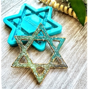 Op de afbeelding: Een Davidster-hanger met een goudkleurig frame en een groen en zilverkleurig interieur. Op de achtergrond een blauwe siliconen mal in de vorm van de Davidster. De hanger ligt op een houten oppervlak.