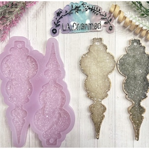 Può includere: Uno stampo in silicone viola chiaro per realizzare decorazioni natalizie, accanto a due decorazioni finite con finiture dorate e interni glitterati. Una decorazione è chiara, l'altra è scura. Un piccolo cartello recita "LA Charmed".
