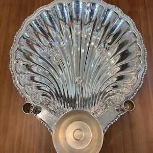 May include: A polished silver-tone metal serving dish shaped like a large scallop shell. The dish features a central circular bowl, two small cylindrical holders on the sides, and a decorative beaded edge along the entire outer rim.
