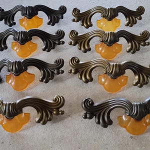 May include: A set of eight drawer pulls featuring an ornate, dark bronze-toned metal frame with scrolling, leaf-like details. Each pull is accented with a translucent, amber-coloured glass or acrylic insert positioned at the centre base.