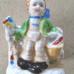 Può includere: Una statuetta in porcellana raffigurante una persona con capelli castano chiaro, che indossa una giacca verde, pantaloni bianchi e scarpe nere. La figura tiene un cestino giallo con un oggetto rosso all'interno su una base bianca.