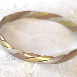 Peut inclure: Un bracelet jonc tricolore composé de trois brins métalliques entrelacés aux tons polis or, argent et cuivre. L'anneau circulaire présente un design torsadé lisse qui crée une apparence tressée continue sur toute la pièce.